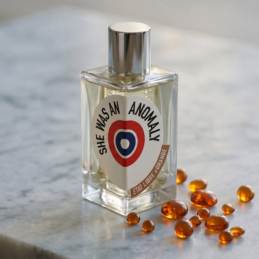 Etat Libre She Was an Anomaly EDP on cream stone with a scatter of honey-colored ambergris flakes
