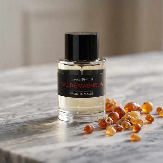 Frederic Malle Eau De Magnolia EDT on pale grey marble with amber resin beads scattered to one side