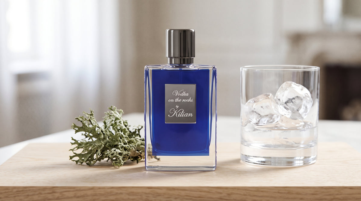 Kilian Vodka On The Rocks EDP on pale sandalwood board with a sprig of oakmoss beside a clear glass tumbler with ice