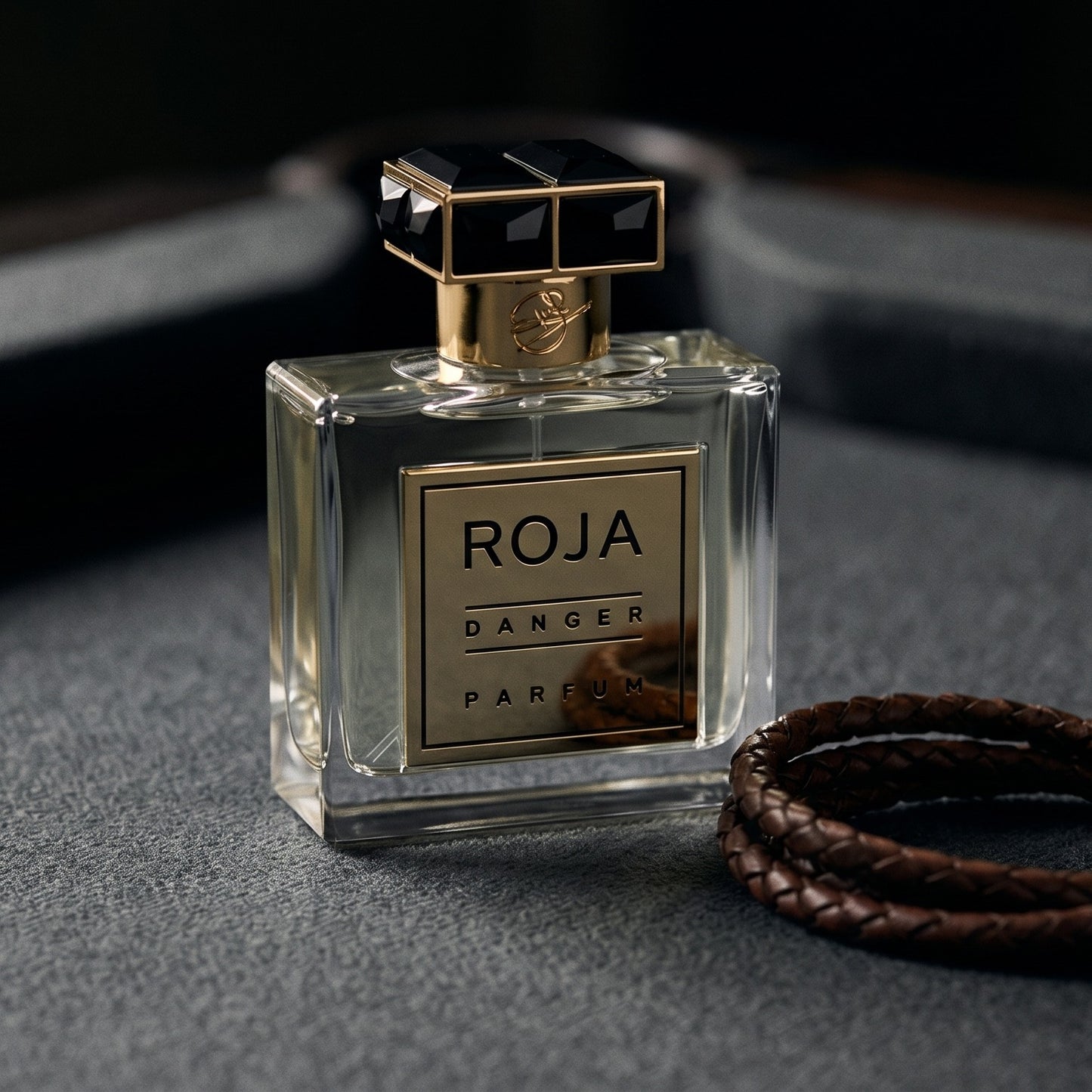Roja Danger Pour Homme Parfum on dark grey baize with a braided leather bracelet coiled loosely