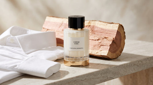 Carolina Herrera Cedar Chic eau de parfum on pale stone beside a folded white cotton shirt and raw cedar wood