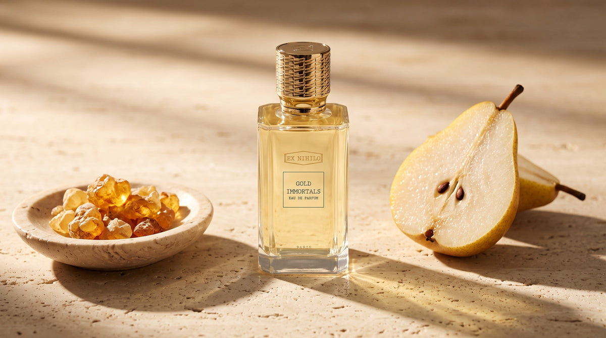 Ex Nihilo Gold Immortals eau de parfum on warm travertine beside a halved pear and golden amber resin