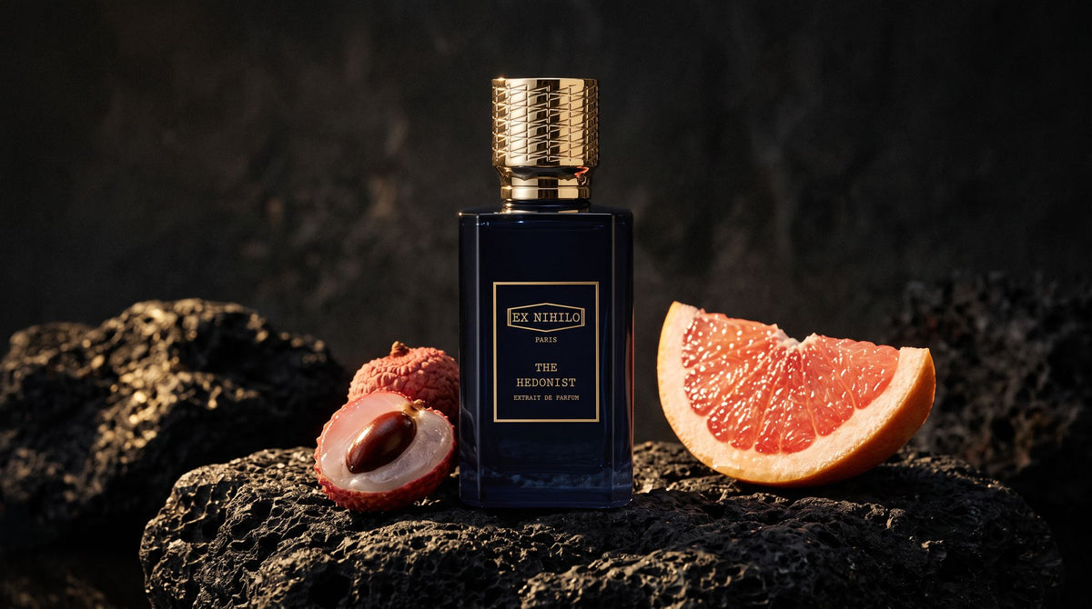 Ex Nihilo The Hedonist Extrait de Parfum bottle on black volcanic stone beside a lychee and a pink grapefruit slice