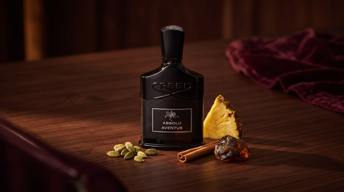 Creed Absolu Aventus black bottle on dark walnut with cardamom pods, a cinnamon stick, pineapple and labdanum resin