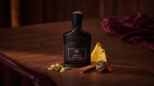 Creed Absolu Aventus black bottle on dark walnut with cardamom pods, a cinnamon stick, pineapple and labdanum resin