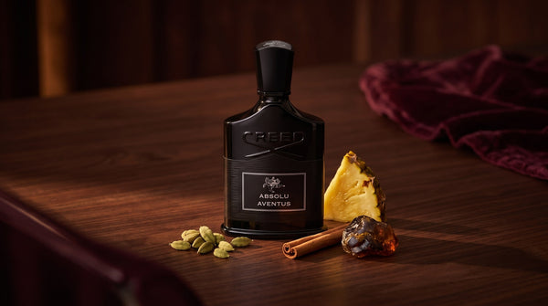 Creed Absolu Aventus black bottle on dark walnut with cardamom pods, a cinnamon stick, pineapple and labdanum resin