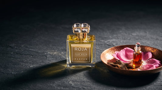 Roja Taif Aoud Parfum on dark stone beside a copper dish of pink damask rose petals, agarwood chips and a small vial of rose oil