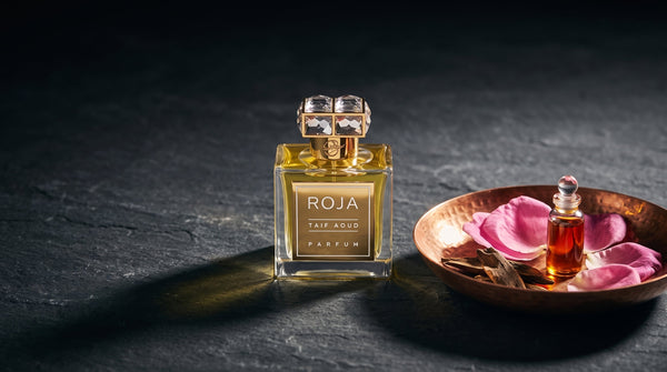 Roja Taif Aoud Parfum on dark stone beside a copper dish of pink damask rose petals, agarwood chips and a small vial of rose oil