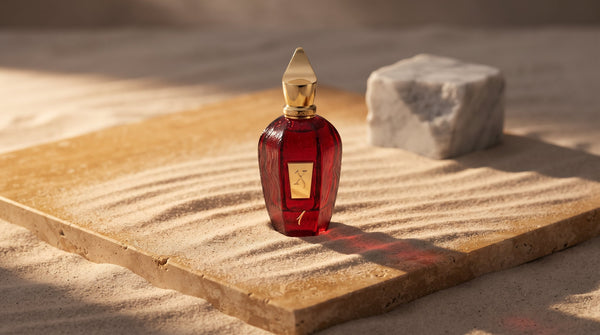 Deep red Xerjoff One flacon with a gold cap on honey travertine stone, fine desert sand drifting across the surface, pale Italian marble behind