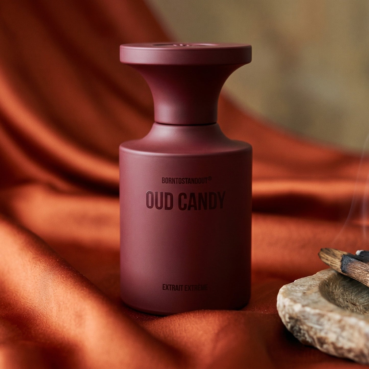 Borntostandout Oud Candy Extrait Extreme on burnt-orange silk with smoldering agarwood on a stone dish