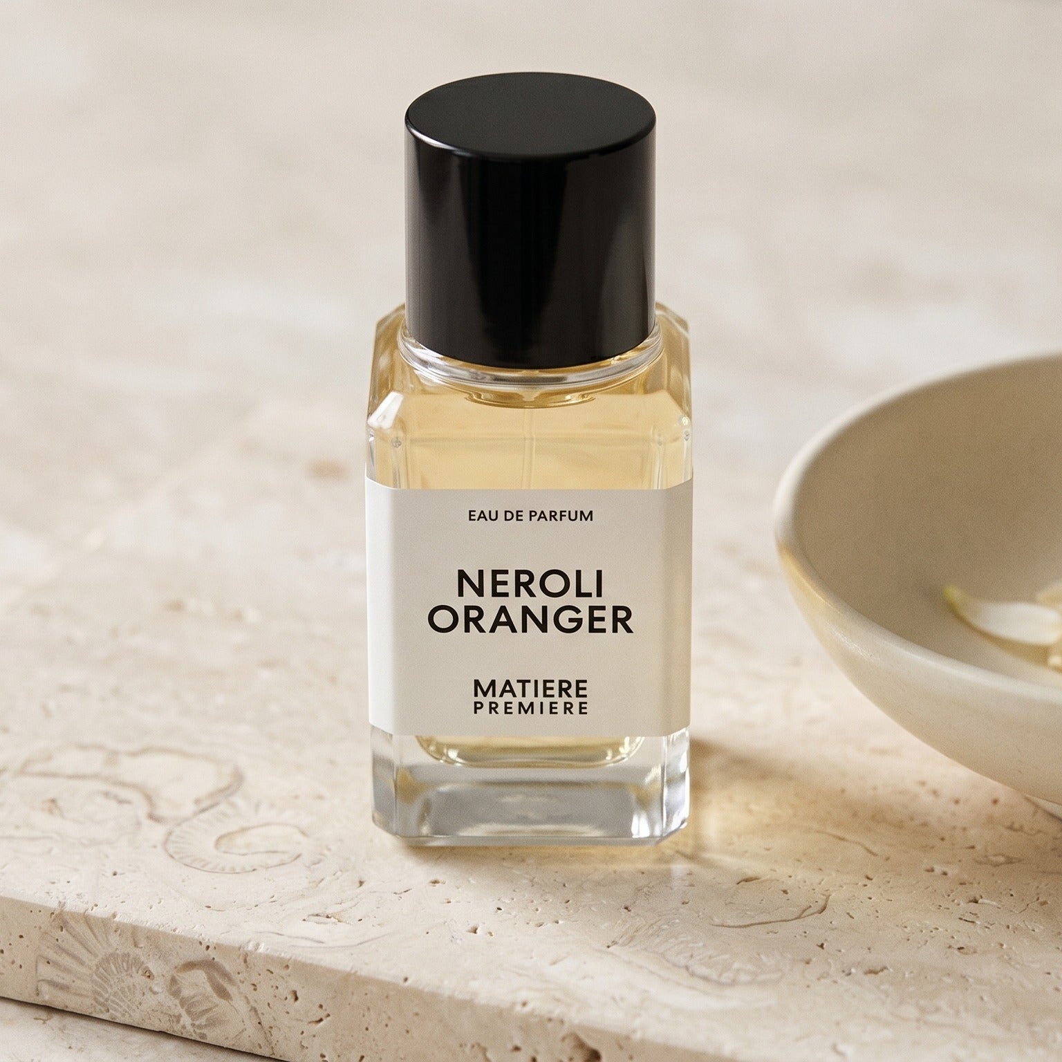 Matiere Premiere Neroli Oranger EDP on pale stone with orange-blossom petals in a shallow bowl