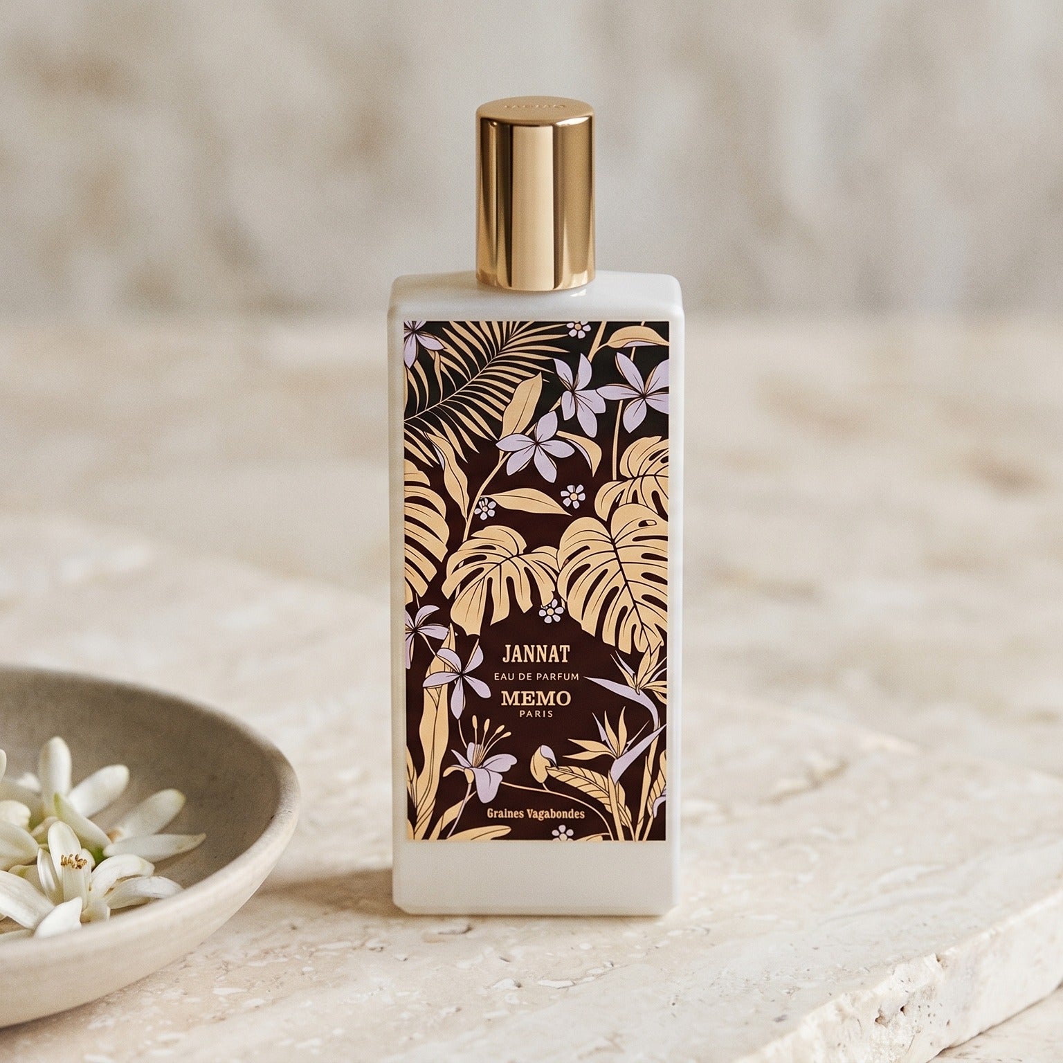 Memo Paris Jannat EDP on pale stone with orange-blossom petals in a shallow bowl