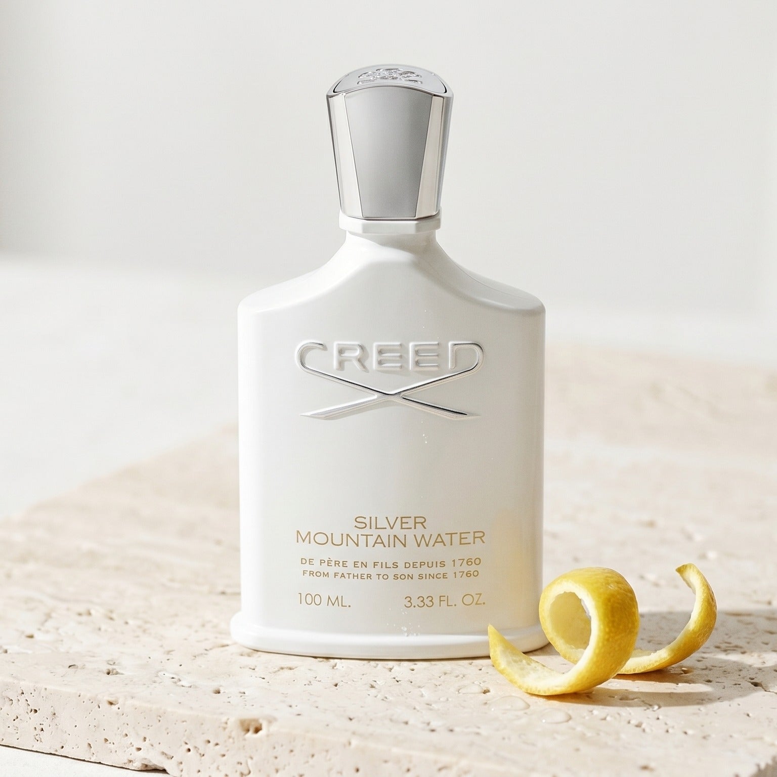 Creed Silver Mountain Water on pale travertine with a lemon peel curled beside the bottle