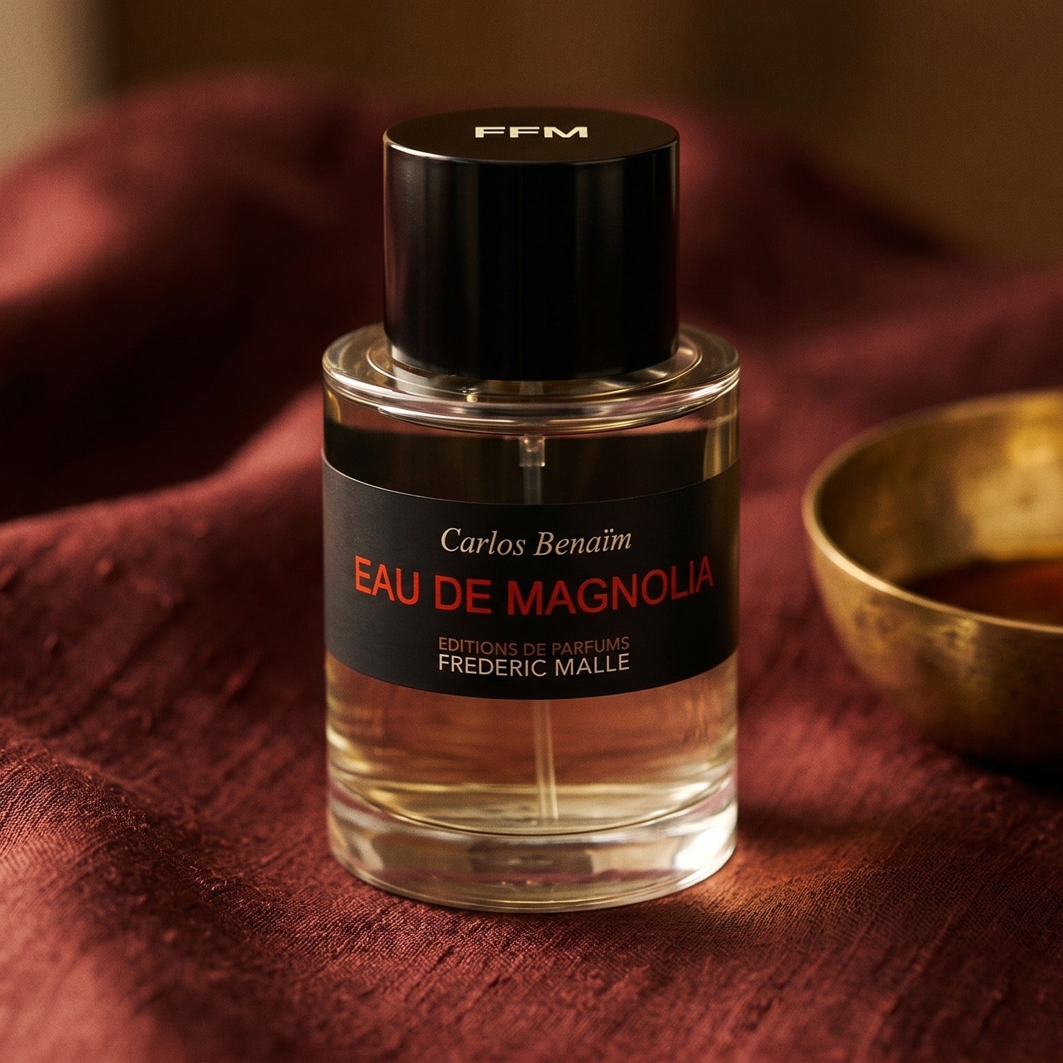 Frederic Malle Eau De Magnolia EDP on amber-toned satin with a gold-rim ceramic dish of labdanum