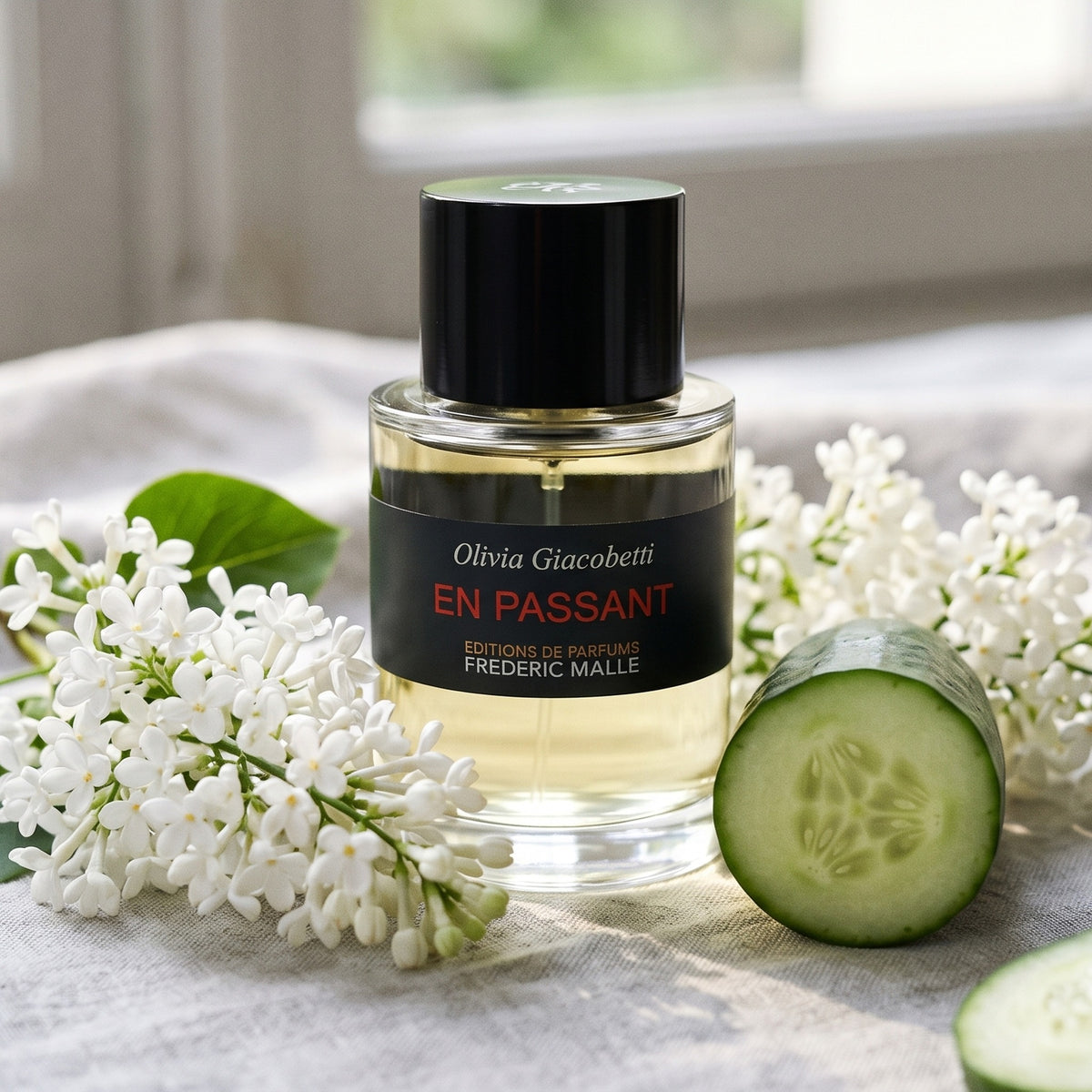 Frederic Malle En Passant EDP on pale grey linen with sprigs of white lilac beside a sliced cucumber