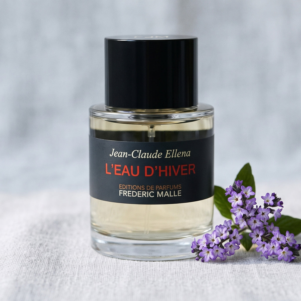 Frederic Malle L'Eau D'Hiver EDT on pale grey linen with a sprig of purple heliotrope
