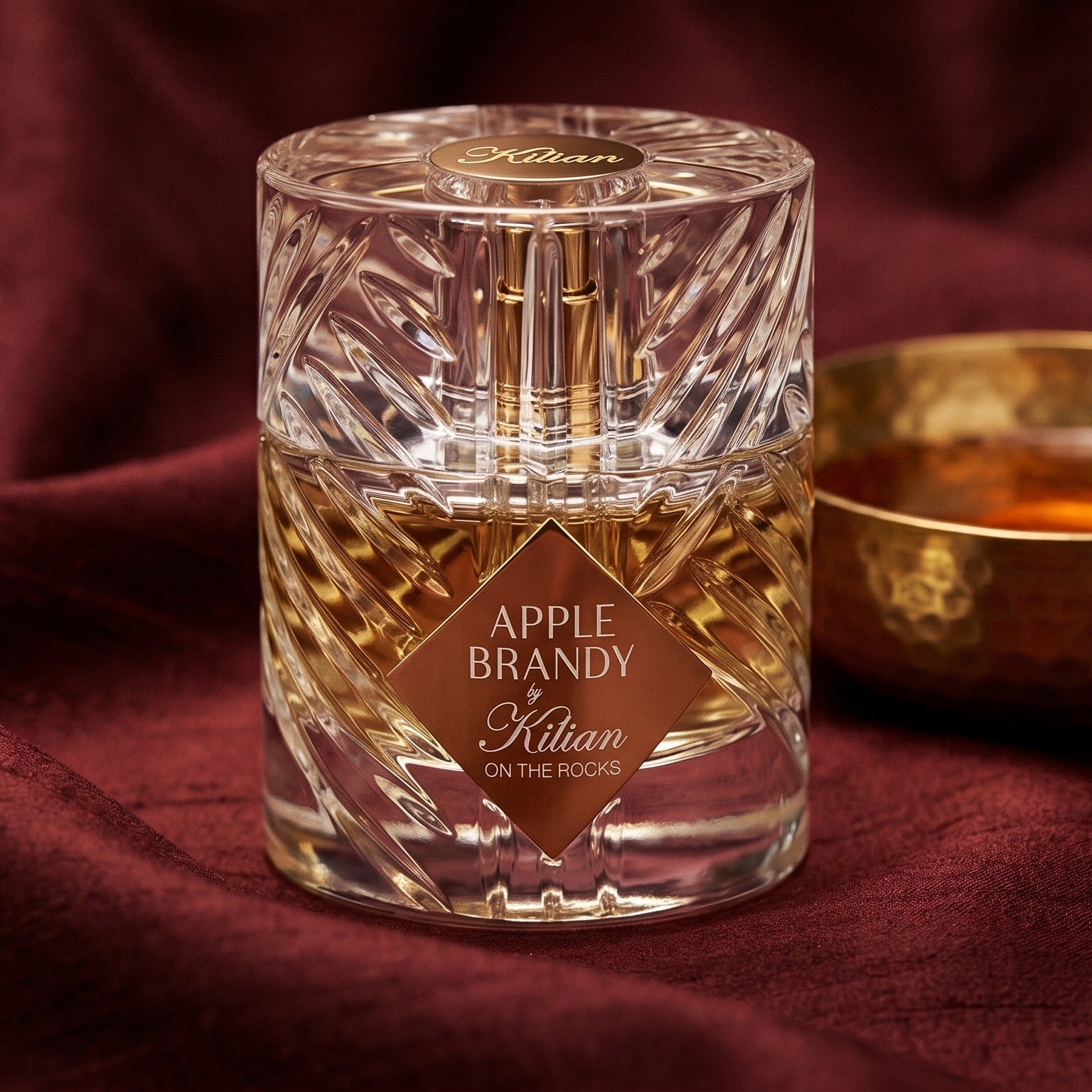Kilian Apple Brandy on the Rocks on russet silk with a matte brass bowl holding tonka and amber pieces