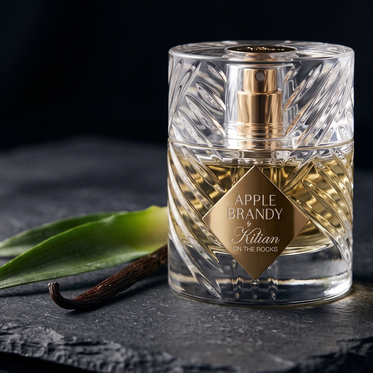 Kilian Apple Brandy on the Rocks on dark slate with a pineapple crown leaf beside a vanilla pod