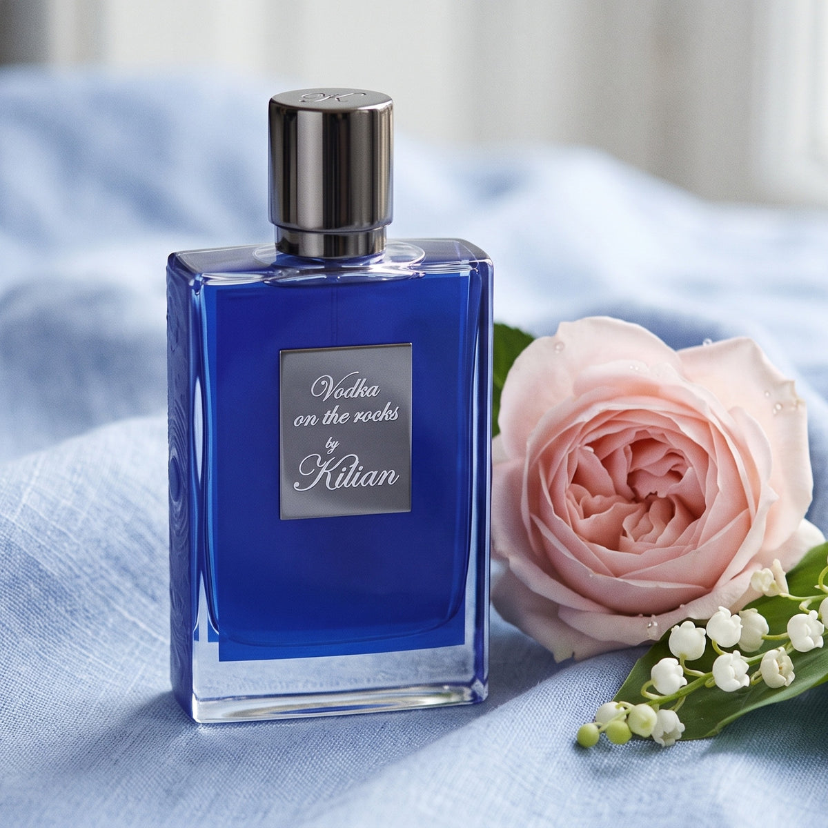 Kilian Vodka On The Rocks EDP on pale blue linen with a sprig of lily of the valley beside a pink rose