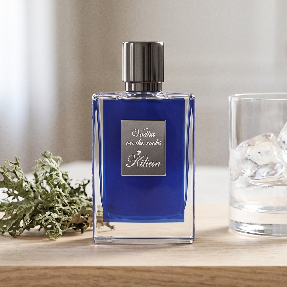 Kilian Vodka On The Rocks EDP on pale sandalwood board with a sprig of oakmoss beside a clear glass tumbler with ice