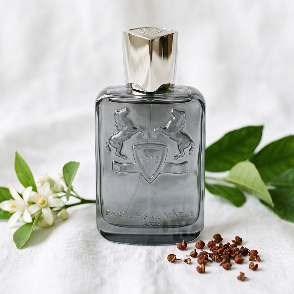 Parfums de Marly Castley EDP on white linen with a sprig of neroli blossom beside lemon-tree leaves and timut peppercorns