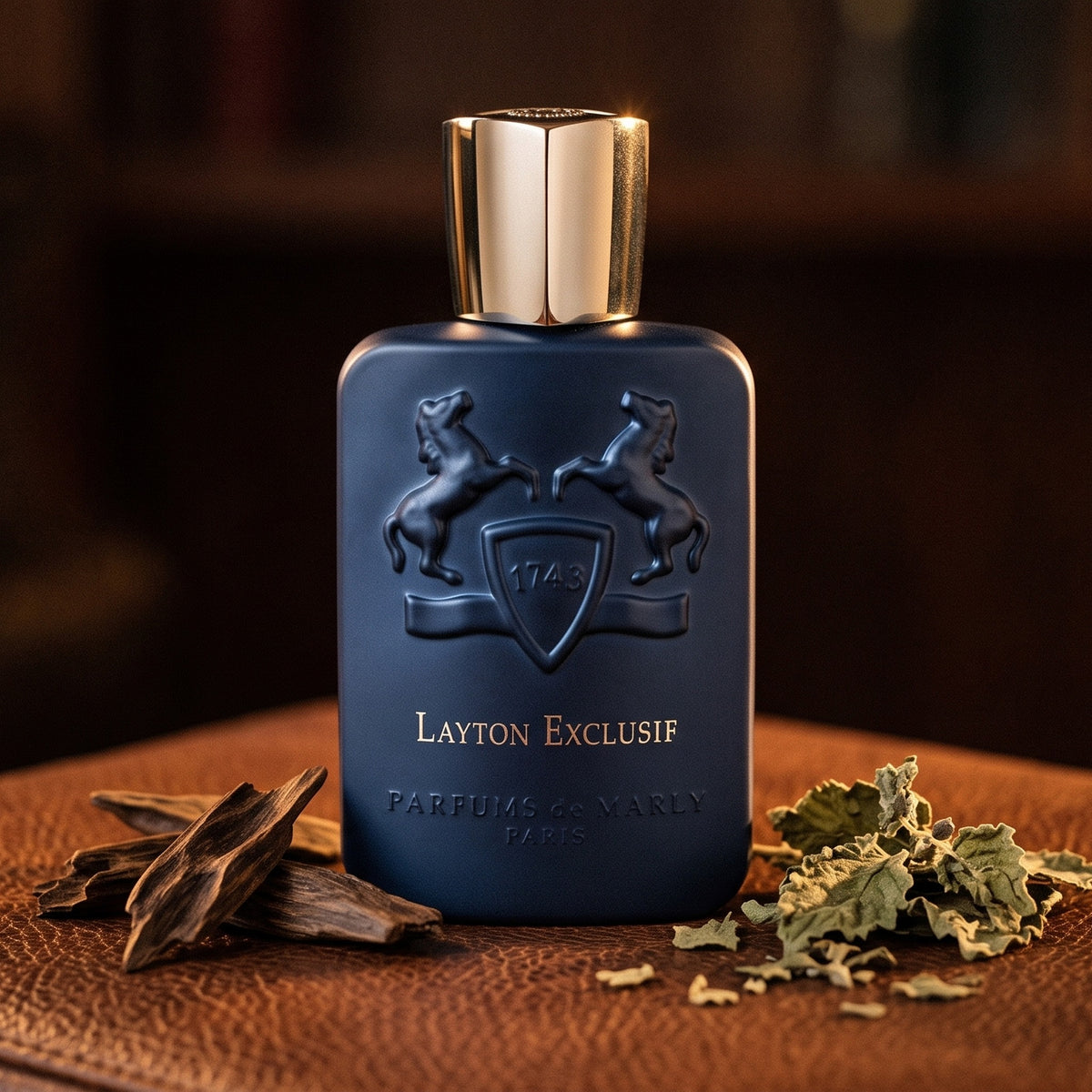 Parfums de Marly Layton Exclusif Parfum on dark tan leather with a few oud wood chips beside dried patchouli leaves