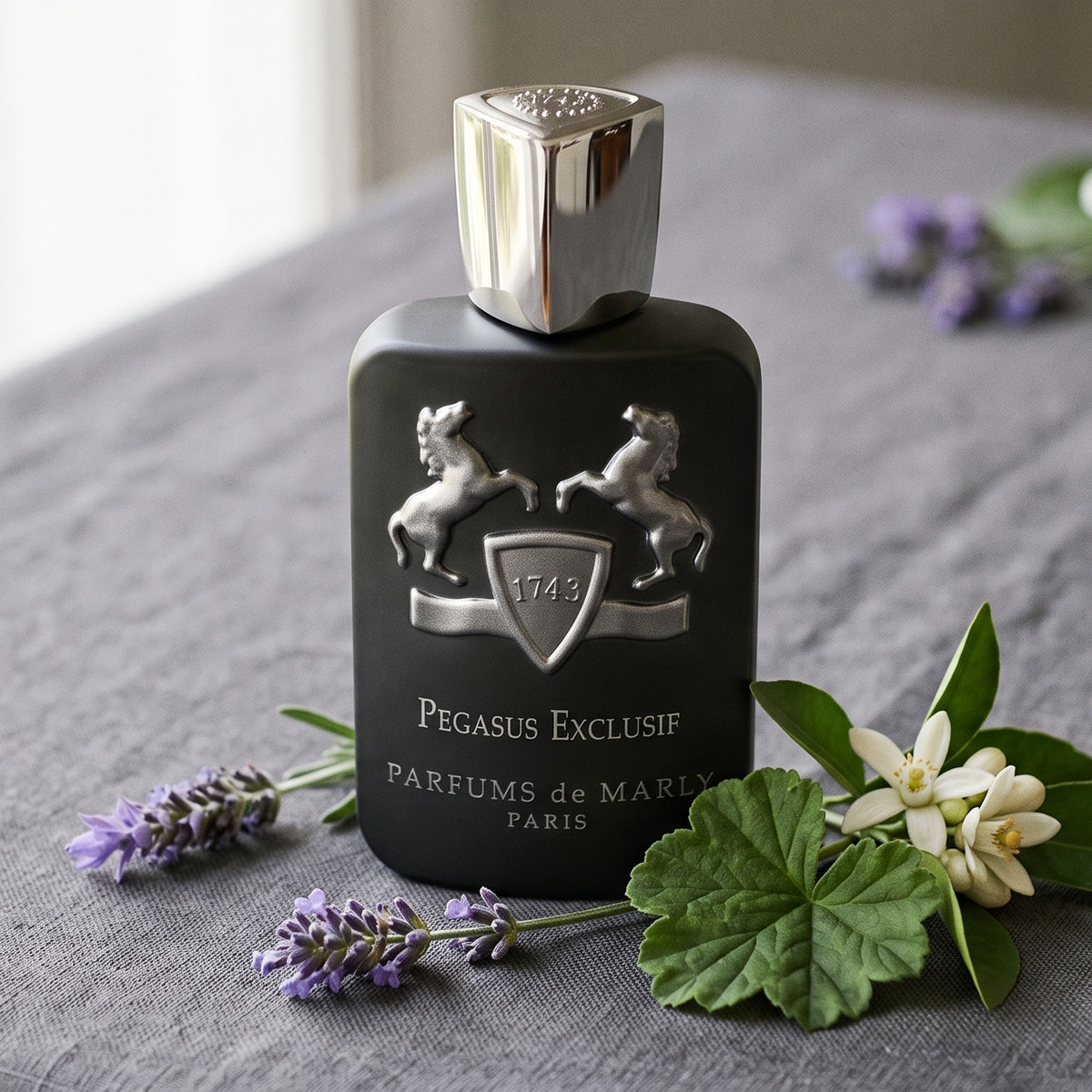 Parfums de Marly Pegasus Exclusif on grey linen with a sprig of lavender beside a geranium leaf and orange blossom