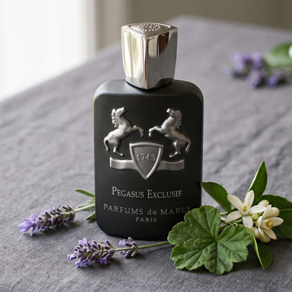 Parfums de Marly Pegasus Exclusif on grey linen with a sprig of lavender beside a geranium leaf and orange blossom