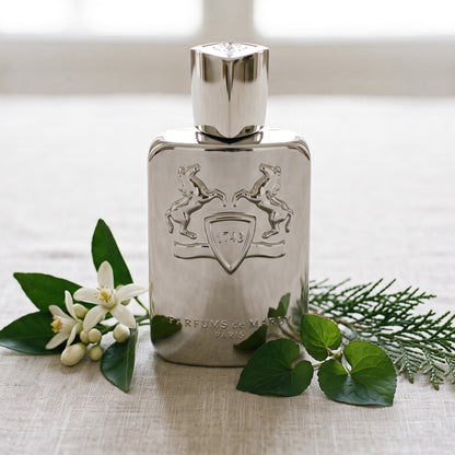 Parfums de Marly Pegasus on pale linen with a sprig of orange blossom beside a few violet leaves and a cypress sprig
