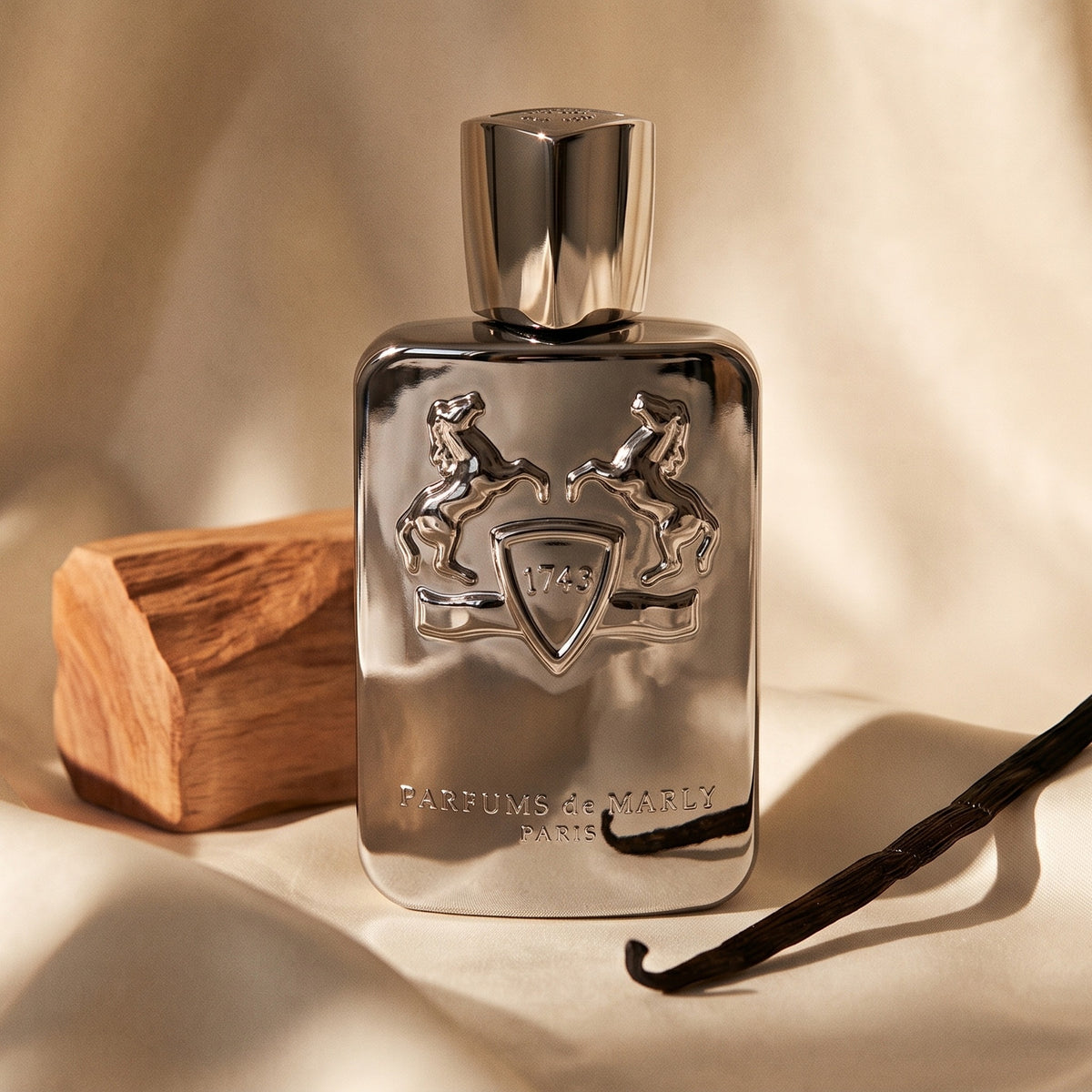 Parfums de Marly Pegasus on cream silk with a vanilla pod beside a small sandalwood block