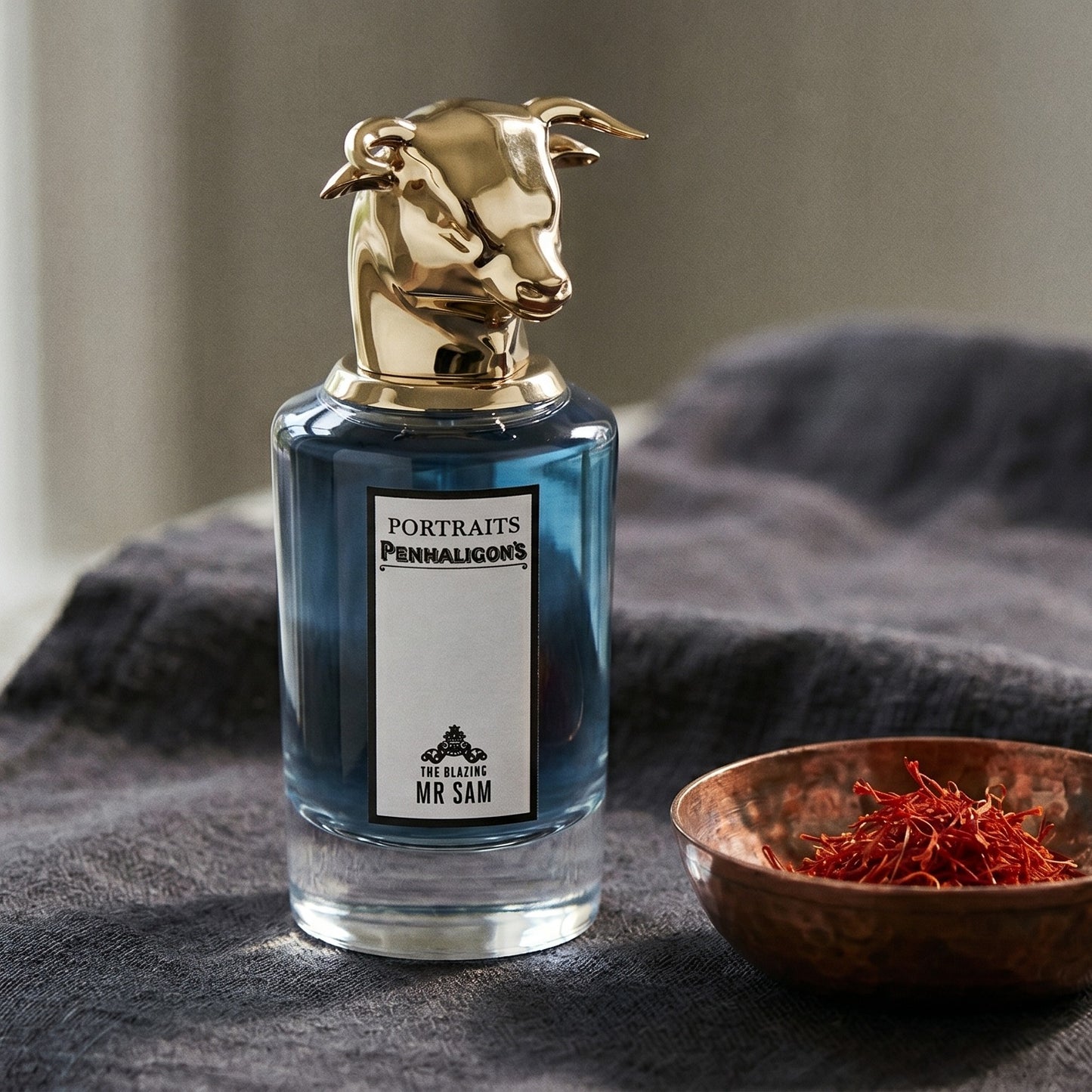 Penhaligon's The Blazing Mister Sam on deep brown linen with a shallow dish of cardamom seeds
