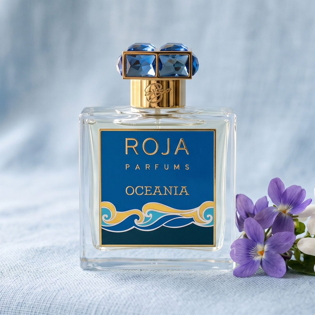 Roja Oceania EDP on pale blue linen with a few violets beside jasmine flowers
