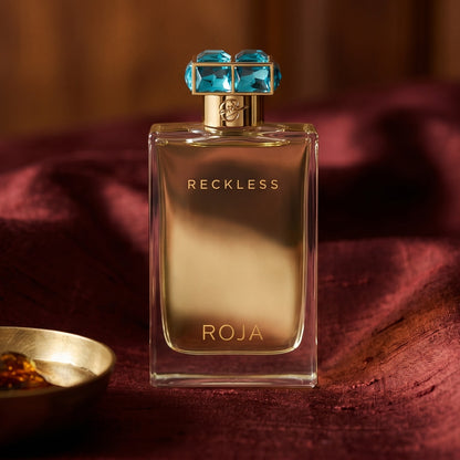 Roja Reckless Pour Femme EDP on amber-toned satin with a gold-rim ceramic dish of labdanum