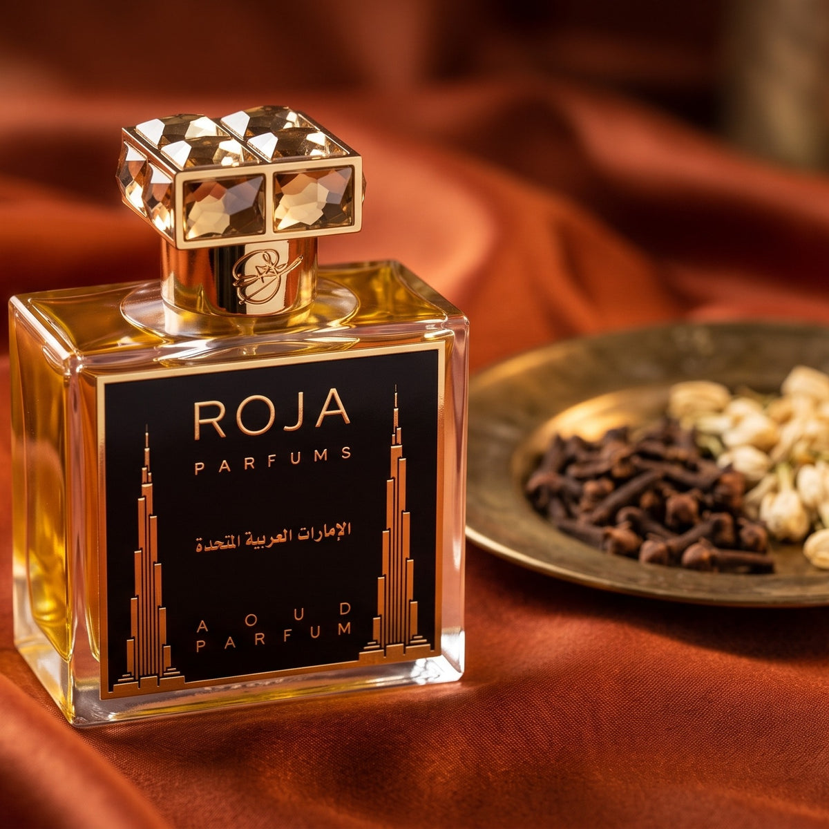 Roja United Arab Emirates Parfum on burnt-orange silk with clove buds and dried jasmine in a small brass saucer